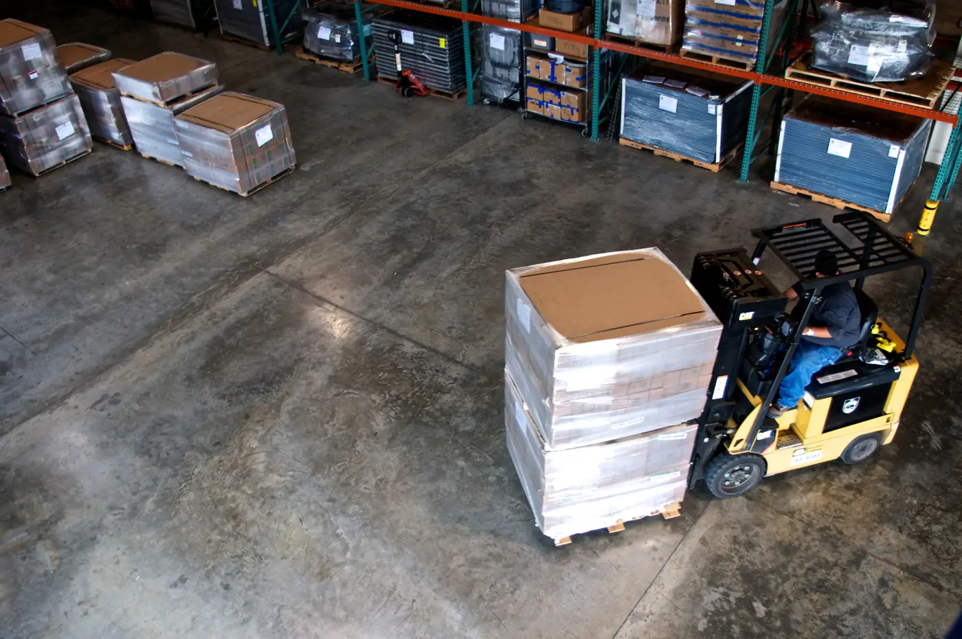 Forklift moving a shrink-wrapped pallet of cartons across a distribution floor lined with pallet racking